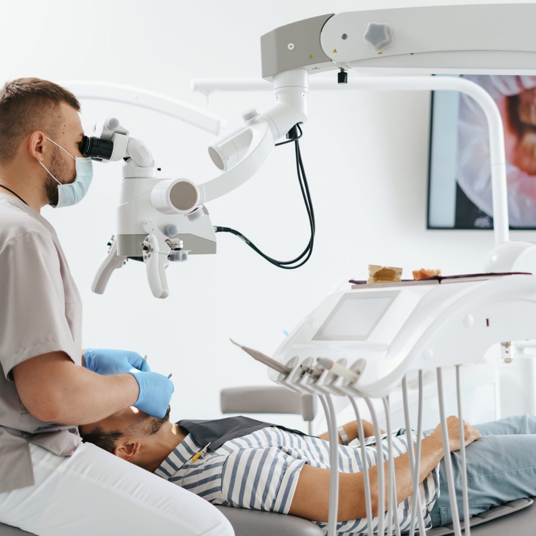 Young man with patient bib on a dental chair and a dentist who sits next to him. He looks on his teeth using a dental microscope and holds a dental bur and a mirror.