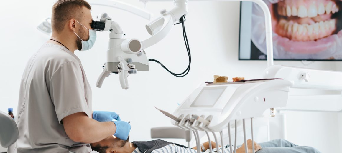 Young man with patient bib on a dental chair and a dentist who sits next to him. He looks on his teeth using a dental microscope and holds a dental bur and a mirror.