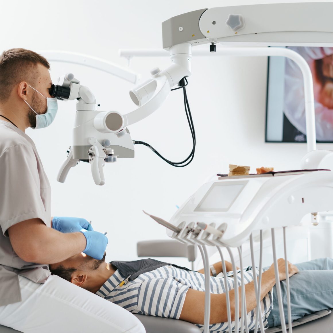 Young man with patient bib on a dental chair and a dentist who sits next to him. He looks on his teeth using a dental microscope and holds a dental bur and a mirror.
