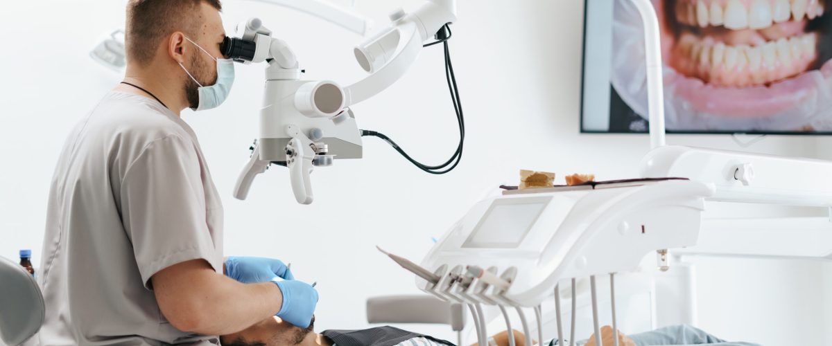 Young man with patient bib on a dental chair and a dentist who sits next to him. He looks on his teeth using a dental microscope and holds a dental bur and a mirror.